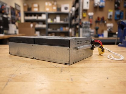 APC APCRBC159 replacement battery cartridge on a wooden workbench, galvanised tray seen from the vented side — AI-generated image
