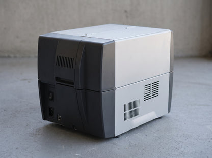 Zebra ZT231 industrial label printer, left rear corner view on a workshop floor — AI-generated image
